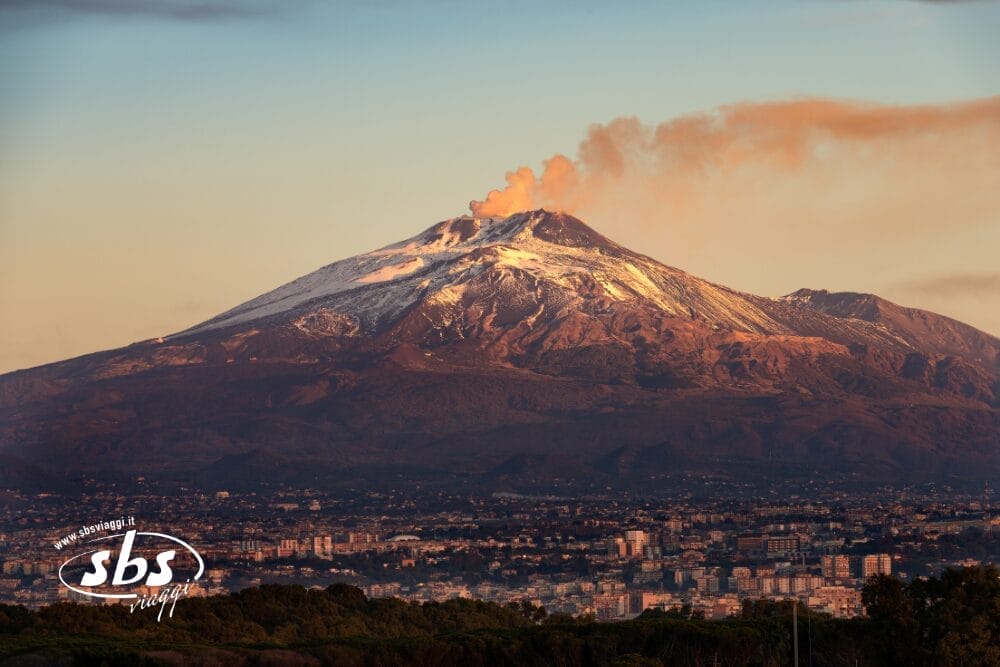 Un vulcano innevato emette fumo sopra una città alla sua base, con nuvole arancioni nel cielo durante il tramonto. Il logo "sbs" e il testo "Bozza automatica" appaiono nell'angolo in basso a sinistra dell'immagine.
