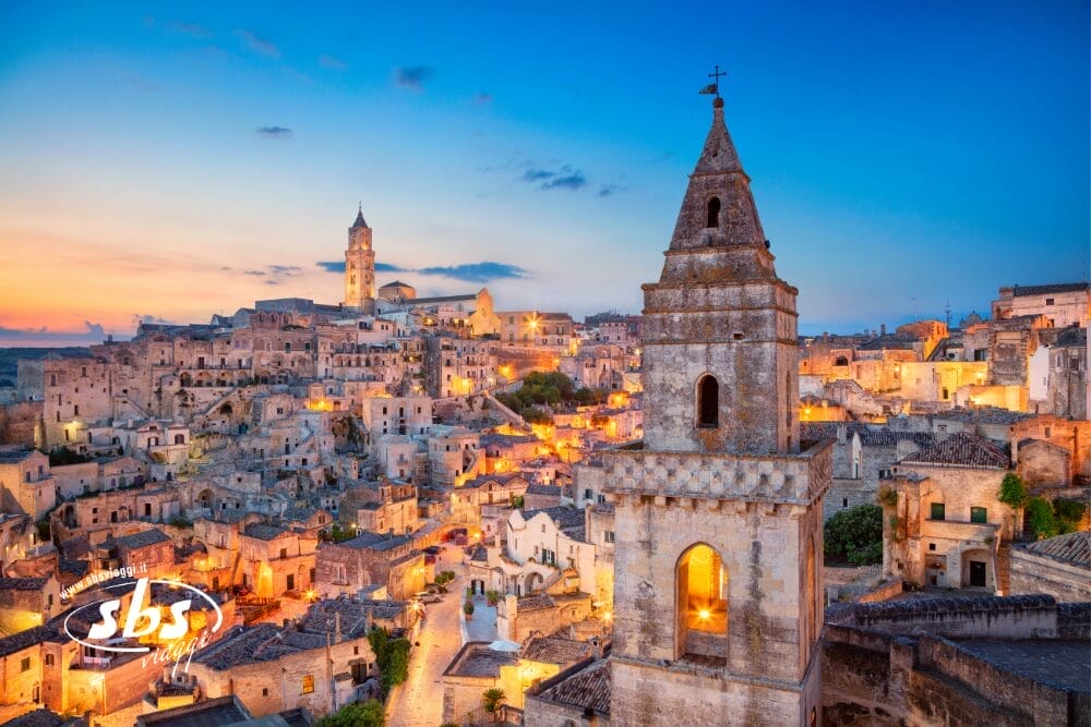 Veduta di Matera, Italia, al tramonto, con antichi edifici in pietra e un importante campanile in primo piano. Le luci calde illuminano la città storica, con la scritta "Bozza automatica" vicino al logo SBS nell'angolo in basso a sinistra.