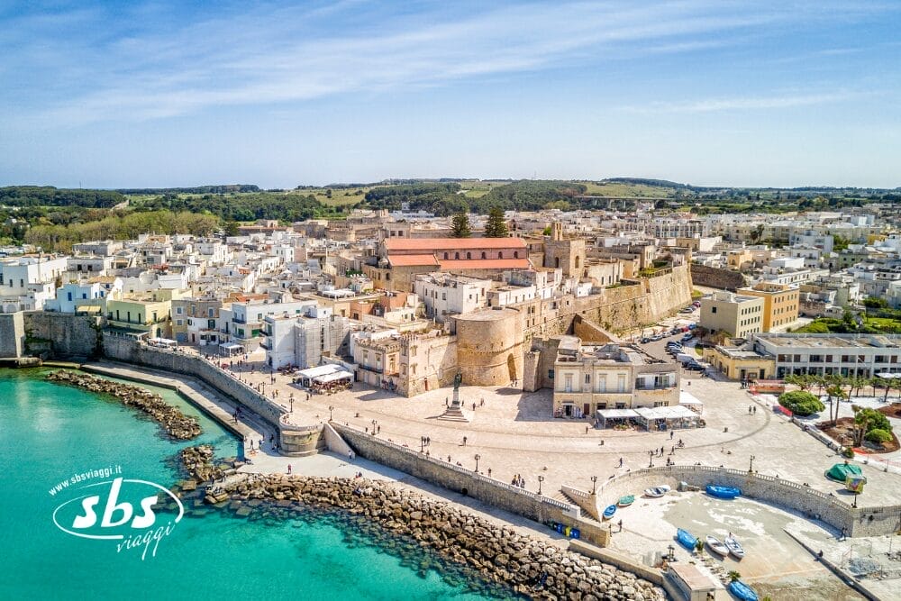 Veduta aerea di una storica città costiera con edifici bianchi, un'importante fortezza, un lungomare in pietra e un mare turchese sotto un cielo limpido. Bozza automatica appare vicino al logo SBS Viaggi nell'angolo in basso a sinistra.