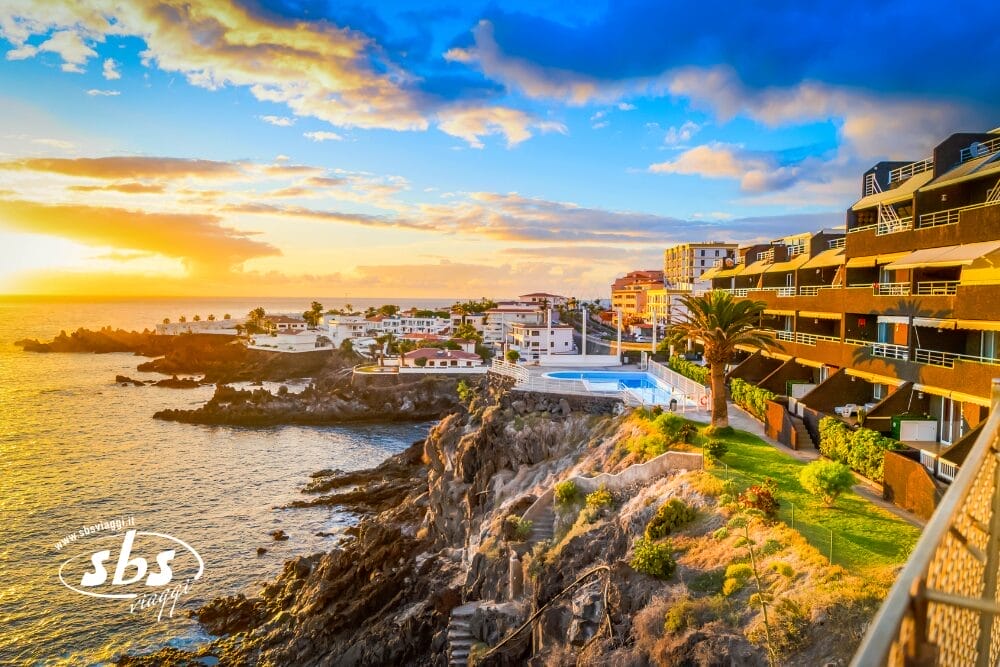 Veduta della costa al tramonto a Tenerife, l'Isola dell'Eterna Primavera, con scogliere rocciose, onde dell'oceano, palme ed edifici moderni con balconi; il cielo parzialmente nuvoloso si illumina di luce calda su questa scena vibrante.
