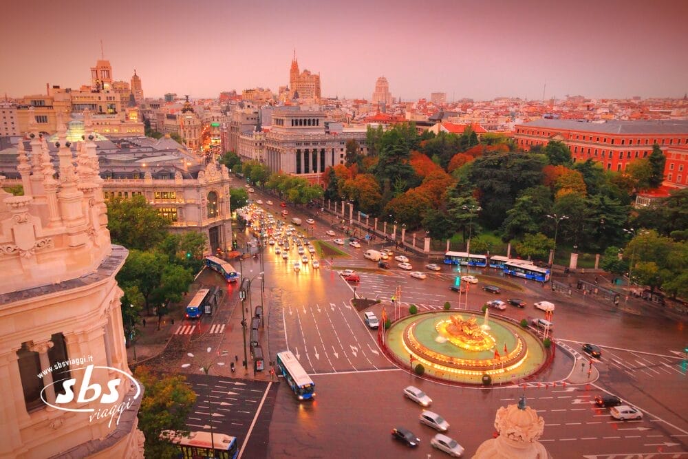 Vista aerea della Plaza de Cibeles di Madrid al tramonto, con la fontana illuminata, le strade trafficate, il verde circostante e gli edifici storici sotto un cielo colorato: una scena perfetta per iniziare il vostro Minitour Castiglia durante la Pasqua.