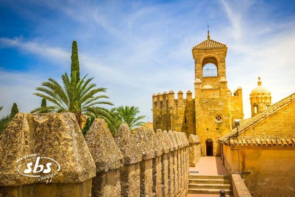 Cammino in pietra con pareti frastagliate che conduce a una torre storica, circondata da palme e cipressi sotto un cielo azzurro. In questa bozza automatica, i dettagli architettonici illuminati dal sole rivelano una sorprendente miscela di stili moreschi ed europei.