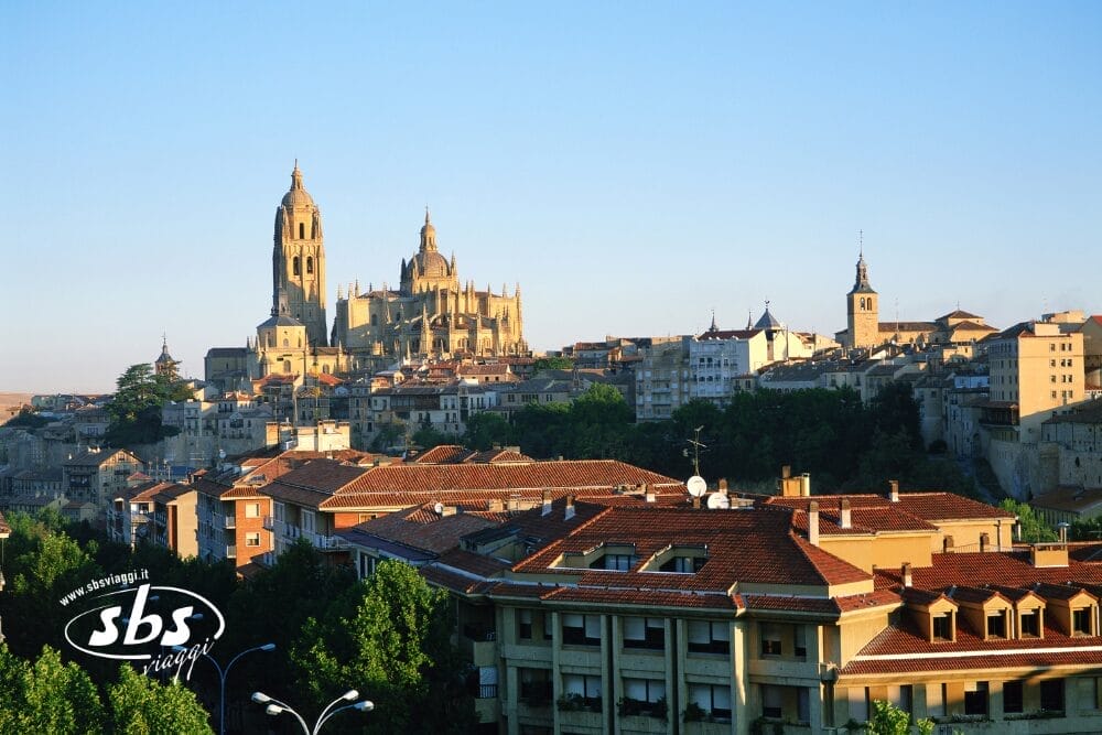 Veduta di Segovia, Spagna, con la maestosa Cattedrale di Segovia con le sue alte guglie che si ergono sopra gli edifici storici e i tetti di tegole rosse sotto un cielo azzurro e limpido. Bozza automatica appare accanto al logo "sbs viajes" nell'angolo in basso a sinistra.