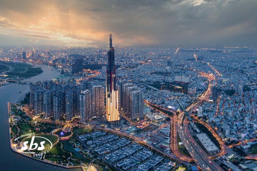 Veduta aerea di una città moderna al tramonto, con un alto grattacielo illuminato circondato da grattacieli e strade tortuose: una vera e propria bozza automatica, mentre le luci della città iniziano a brillare sotto un cielo drammatico lungo il fiume.