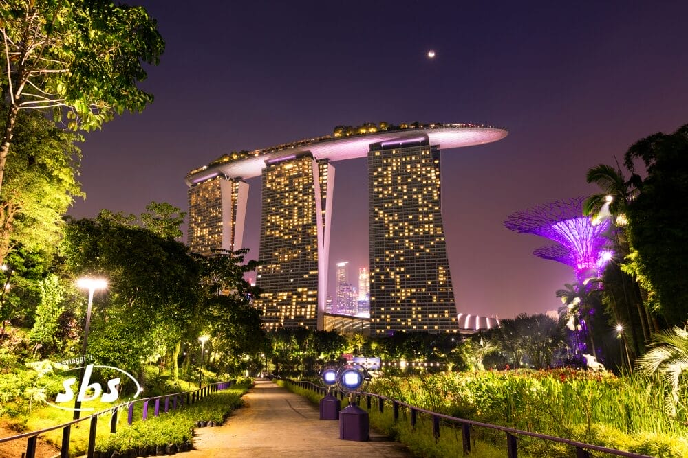 Vista notturna dell'hotel Marina Bay Sands di Singapore, illuminato dalle luci. Il cielo è viola e anche le vicine strutture Supertree e i giardini lussureggianti sono illuminati, creando una scena incantevole come quella di Bali o Gili Trawangan. Un sentiero si snoda nel verde.