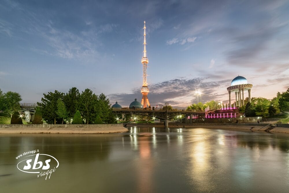 Un fiume che riflette le luci della Tashkent TV Tower illuminata e degli edifici vicini al tramonto, con alberi, un padiglione blu a cupola sulla destra e il logo di SBS Viaggi nell'angolo in basso a sinistra. Bozza automatica appare sottilmente nella composizione.