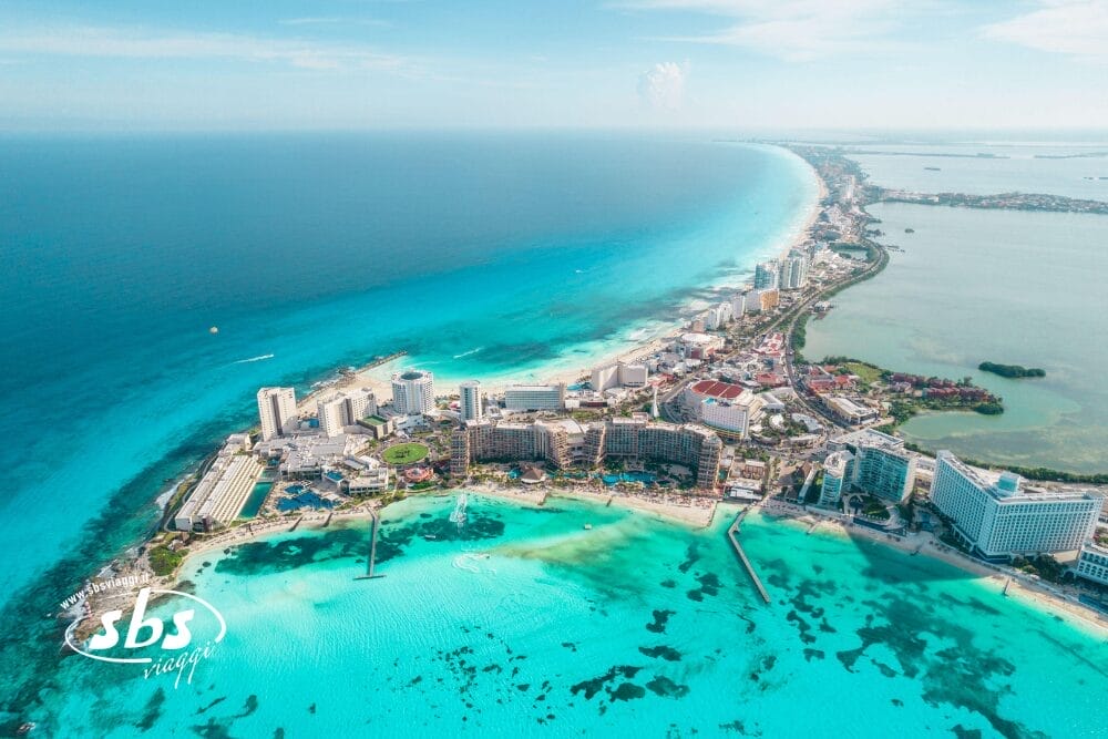 Vista aerea della zona alberghiera di Cancun su un Tour Messico, con alti resort lungo una stretta striscia tra l'oceano turchese e la laguna, sotto un cielo azzurro e limpido.