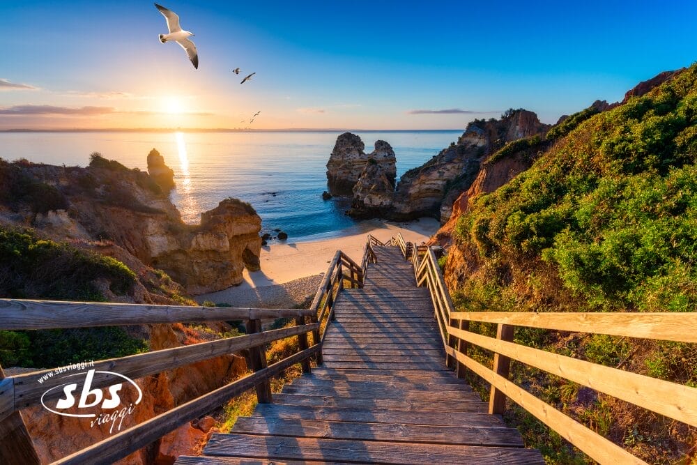 Scale di legno conducono a una spiaggia sabbiosa circondata da scogliere rocciose al sorgere del sole in Algarve, nel Sud del Portogallo. Il sole sorge sulle acque calme dell'oceano, con i gabbiani che volano sopra e la vegetazione lussureggiante sulla destra.