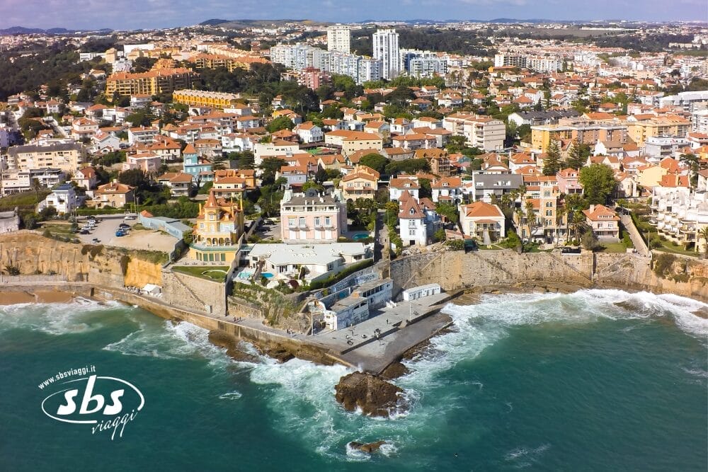 Veduta aerea di una città costiera con edifici dai tetti arancioni, un litorale roccioso e onde che si infrangono sulla riva. Il logo di SBS Viaggi e la scritta "Bozza automatica" appaiono nell'angolo in basso a sinistra.