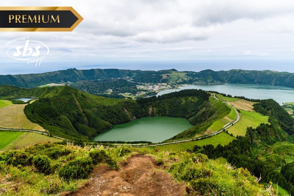 Una vista panoramica di colline verdi, un lago craterico e un villaggio lontano sotto un cielo nuvoloso, con "PREMIUM", "Bozza automatica" e un logo bianco in sovrimpressione nell'angolo superiore sinistro.
