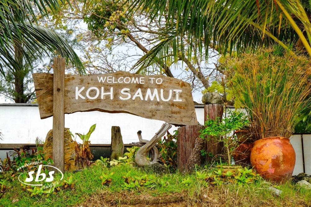 Un'insegna rustica in legno recita "Welcome to Koh Samui", circondata da una vegetazione lussureggiante, palme, una Bozza automatica e un grande vaso di terracotta, creando una scena tropicale e invitante.