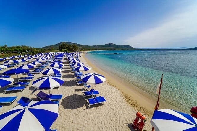 Una spiaggia sabbiosa fiancheggiata da file di ombrelloni blu e bianchi e sedie a sdraio abbinate, accanto a un'acqua turchese e trasparente, con colline verdi sullo sfondo sotto un cielo azzurro brillante: un luogo perfetto per rilassarsi o per abbozzare la prossima Bozza automatica.