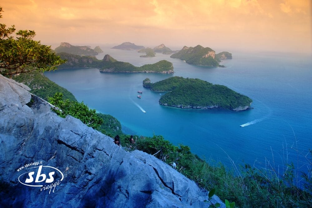 Vista da una scogliera rocciosa che si affaccia su un mare azzurro punteggiato di isole verdi e lussureggianti, con barche che lasciano scie bianche nell'acqua sotto un cielo arancione parzialmente nuvoloso al tramonto. Il logo "sbs viaggi" e Bozza automatica appaiono nell'angolo in basso a sinistra.