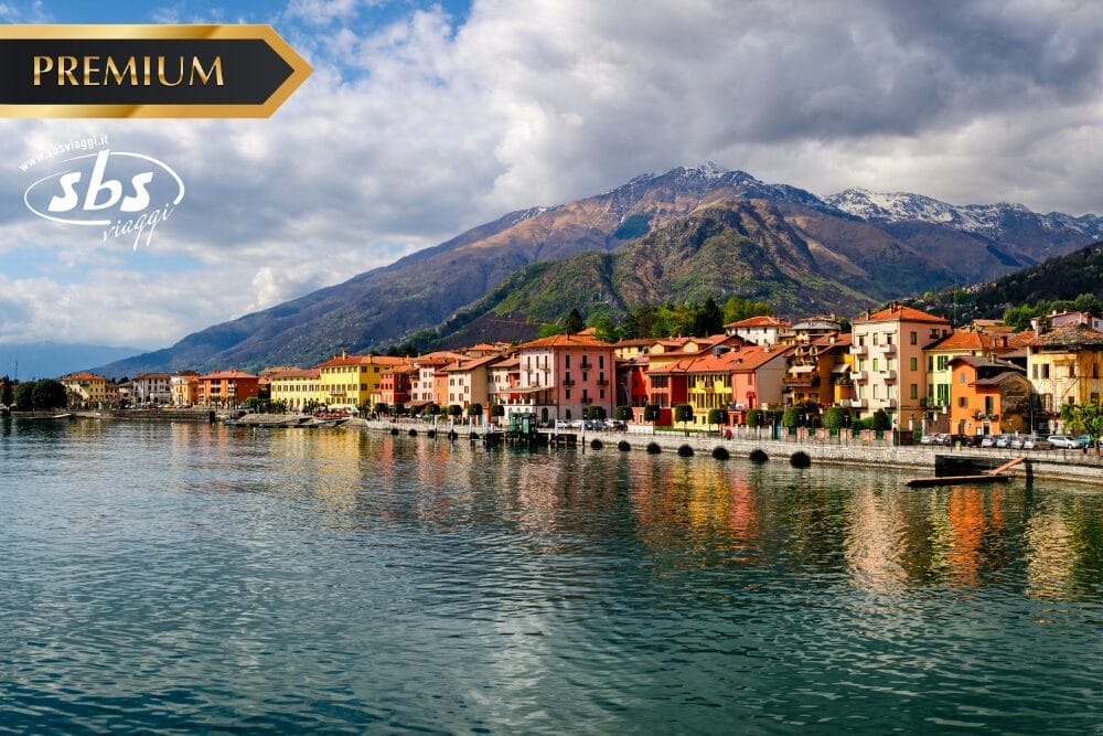 Edifici colorati costeggiano un lungomare con le montagne sullo sfondo, sotto un cielo parzialmente nuvoloso. L'acqua calma riflette le case vivaci. I loghi "PREMIUM", "sbs viaggi" e Bozza automatica appaiono nell'angolo in alto a sinistra.