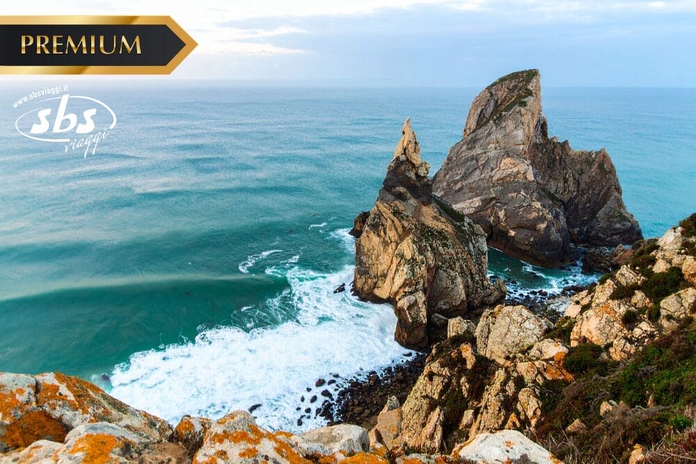 Scogliere rocciose che si affacciano sulle onde turchesi dell'oceano, con una spettacolare formazione rocciosa che emerge dall'acqua. I loghi "PREMIUM" e "sbs voyages" appaiono nell'angolo in alto a sinistra, insieme a una sottile filigrana automatica Bozza.