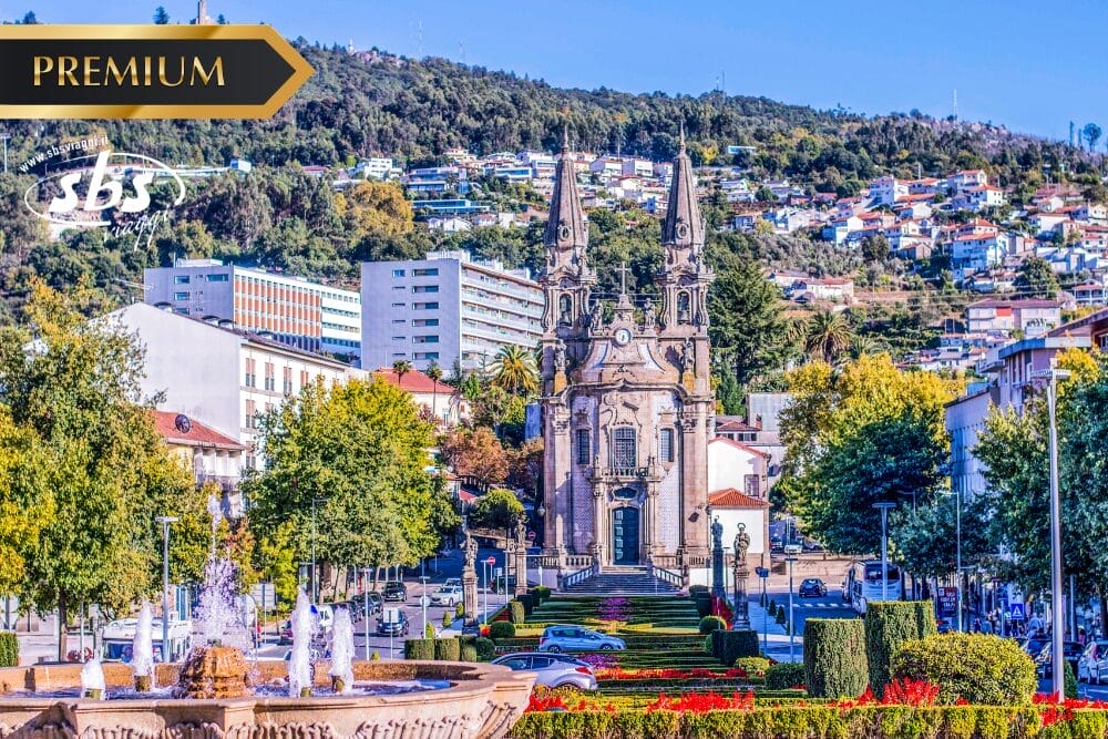 Una grande chiesa con due guglie si trova al centro di un viale con giardino, circondata da edifici colorati e colline verdi; una fontana, come un capolavoro di bozza automatica, è in primo piano sotto un cielo azzurro e limpido.