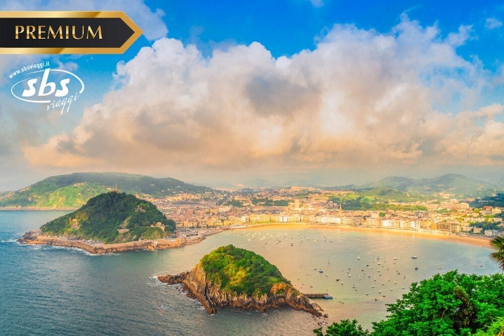 Veduta aerea di una città costiera della Spagna del Nord con colline verdeggianti, spiagge sabbiose e una baia piena di barche sotto un cielo parzialmente nuvoloso. Nell'angolo in alto a sinistra compaiono l'etichetta "PREMIUM" e il logo "sbs viaggi".