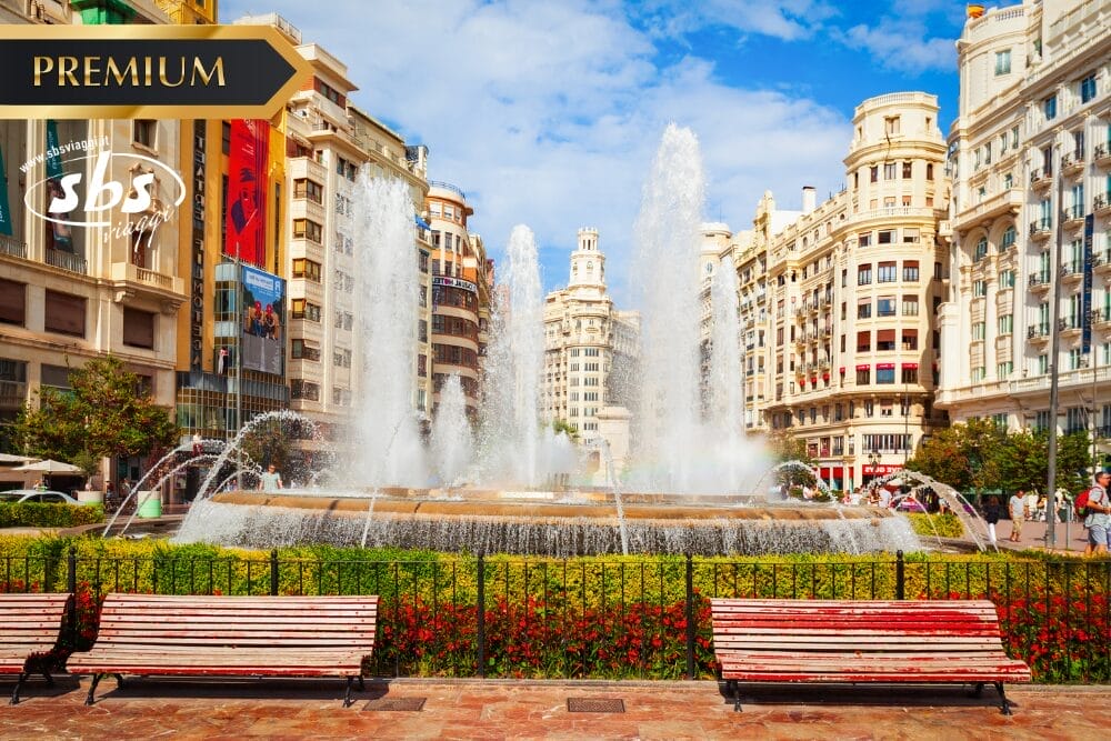 Una grande fontana spruzza acqua al centro di una vivace piazza cittadina, circondata da edifici storici ornati sotto un cielo azzurro. Due panchine vuote si trovano in primo piano, mentre fiori e verde costeggiano l'area della fontana automatica Bozza.