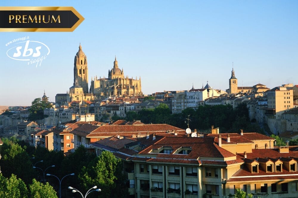 Una veduta panoramica di una città storica con una grande cattedrale e antichi edifici, circondata da alberi sotto un cielo azzurro e limpido. I loghi "PREMIUM", "sbs viaggi" e "Bozza automatica" sono sovrapposti negli angoli superiori.