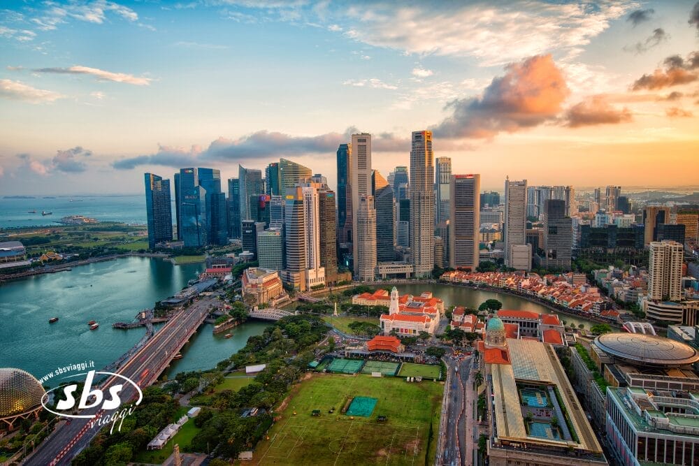 Vista aerea del moderno skyline di Singapore al tramonto, con alti grattacieli e Marina Bay. In primo piano gli edifici dai tetti rossi del quartiere storico e la Bozza automatica, mentre la calda luce del sole illumina il cielo parzialmente nuvoloso.