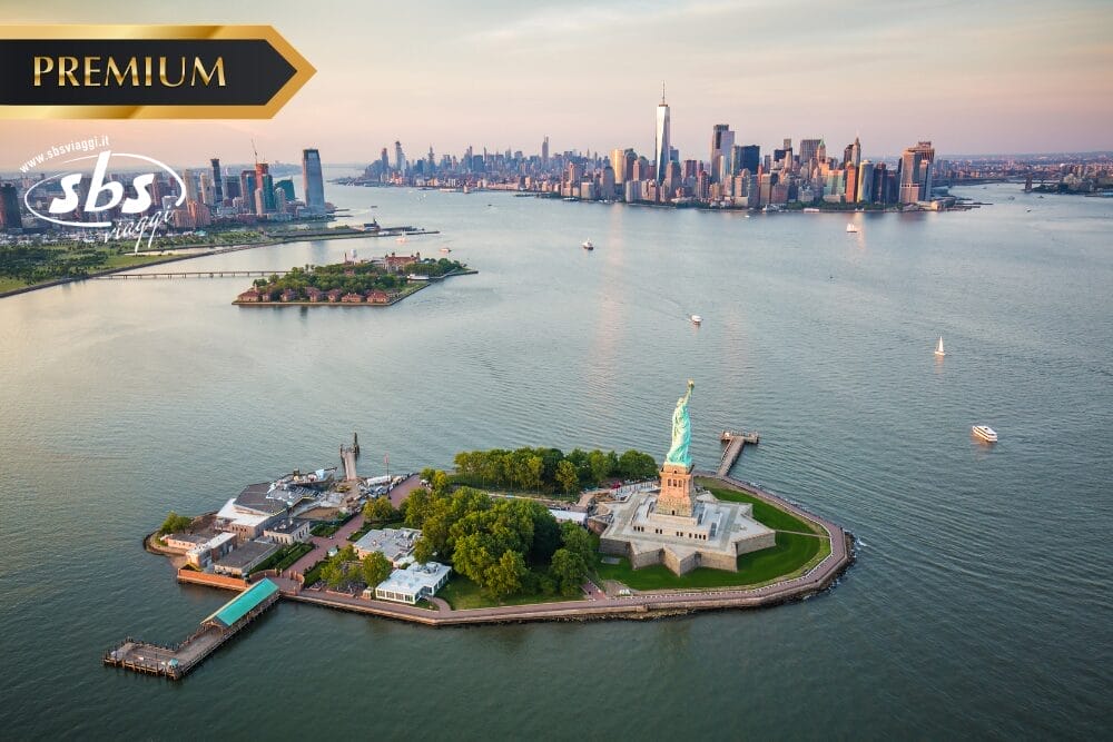 Veduta aerea della Statua della Libertà su Liberty Island con lo skyline di Manhattan al tramonto. Nell'acqua sono visibili delle barche e un'etichetta "Bozza automatica". I loghi "PREMIUM" e "sbs" sono in alto a sinistra.