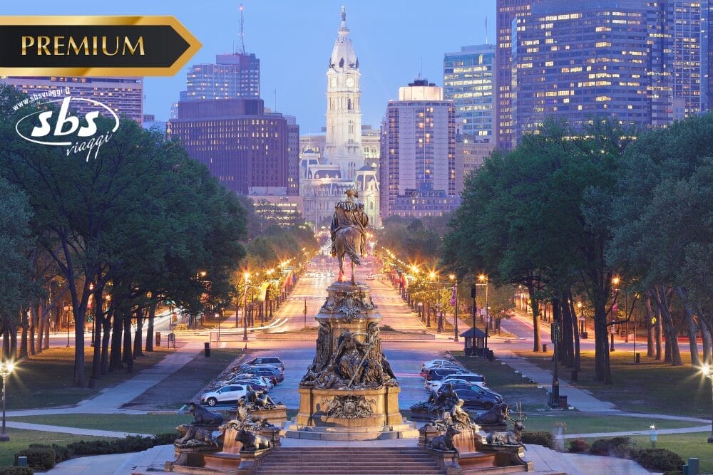 Una veduta della città di Filadelfia al tramonto, con il viale alberato Benjamin Franklin Parkway, la statua Eakins Oval e i grattacieli illuminati con al centro il Municipio, che cattura lo spirito di città come New York. Sono visibili i loghi "PREMIUM" e "sbs viaggi".