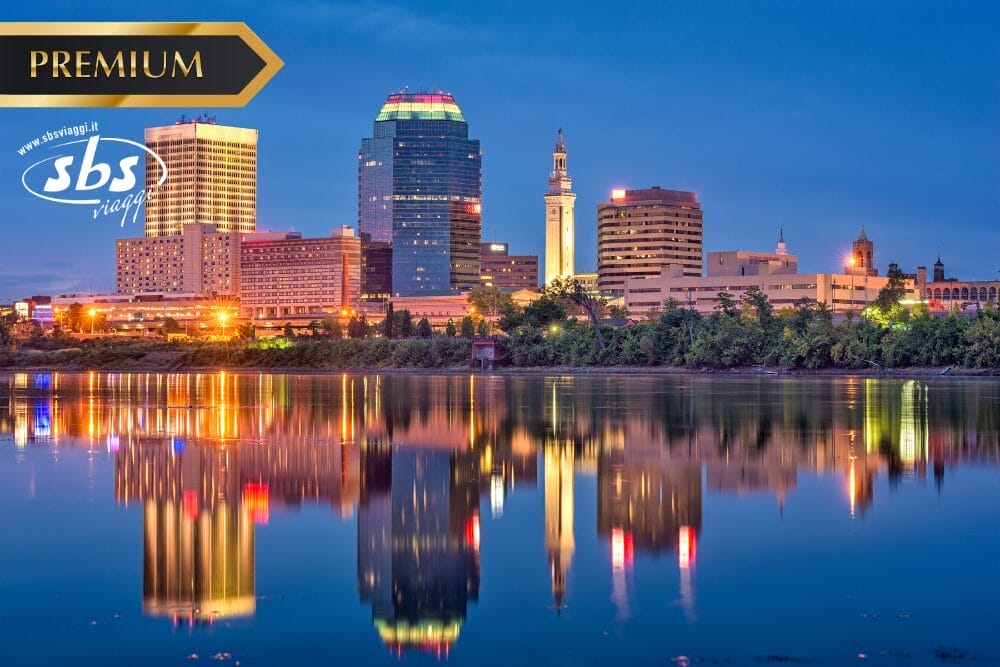 Lo skyline della città al tramonto si riflette in un fiume calmo, con edifici illuminati e una torre prominente. I loghi "PREMIUM", "sbs viaggi" e "Bozza automatica" appaiono nell'angolo in alto a sinistra.