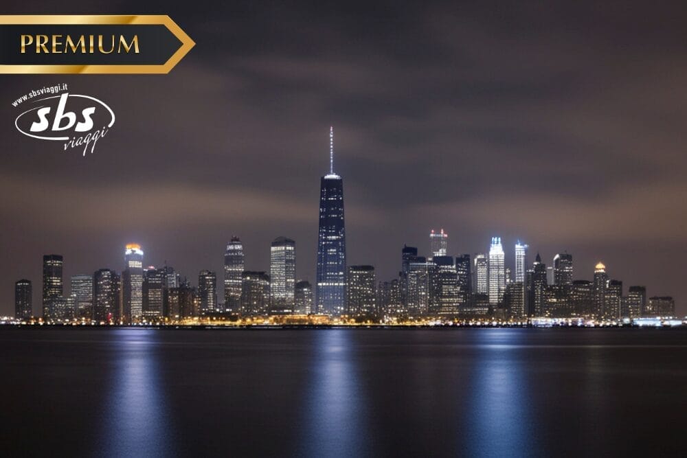Veduta notturna dello skyline di una città con alti edifici riflessi nell'acqua calma; l'edificio più alto è centrato. L'immagine presenta un banner "PREMIUM", il logo "sbs viaggi" e un'etichetta Bozza automatica nell'angolo in alto a sinistra.