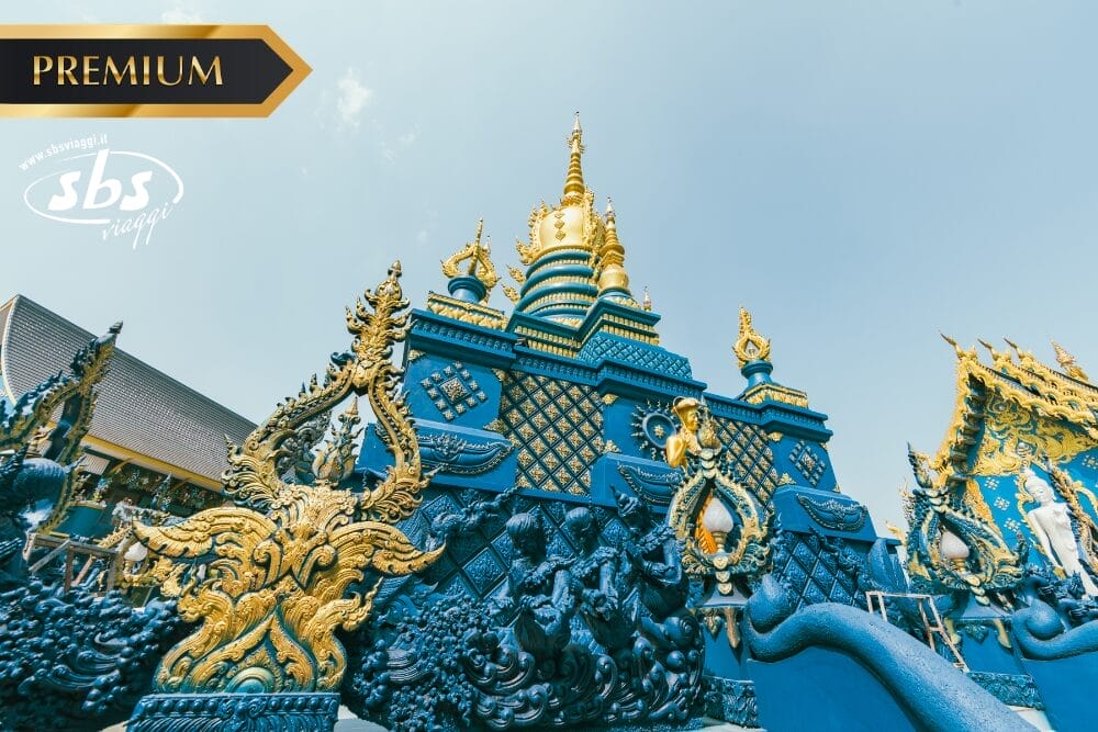Tempio ornato di blu e oro con intricate sculture di draghi, guglie dorate e dettagli decorativi sotto un cielo limpido a Chiang Mai. I loghi "Premium" e "sbs" sono visualizzati sull'immagine.