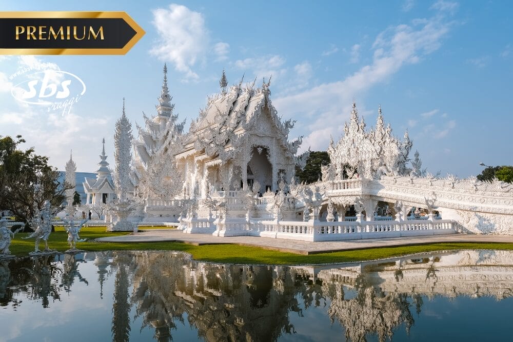 Uno splendido tempio bianco con dettagli intricati si riflette in uno stagno sotto un cielo blu. Il tempio, che fa parte del Thailandia Gran Tour, presenta sculture e guglie decorate. Il tempio è circondato da un'erba verde, con un'etichetta "PREMIUM" nell'angolo in alto a sinistra.