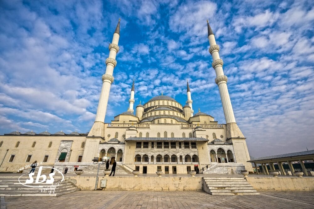 Grande moschea con quattro alti minareti, finestre ad arco e tetti a cupola contro un cielo luminoso e parzialmente nuvoloso; alcune persone sono in piedi su ampi gradini di pietra di fronte, catturando la scena come un Bozza automatico di bellezza architettonica.