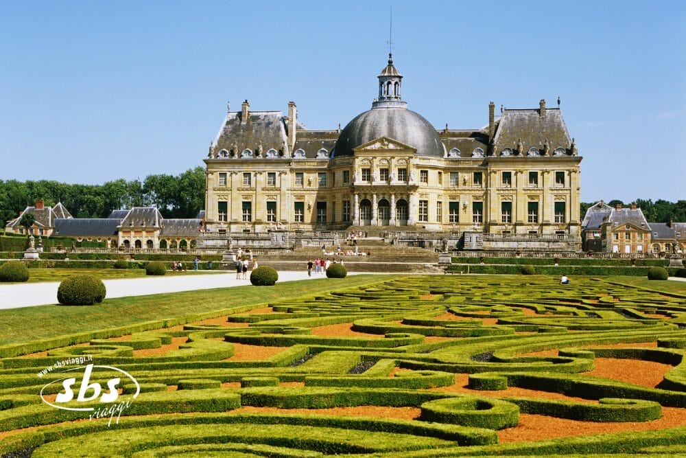 Un grande castello con tetto centrale a cupola si erge dietro giardini ornati e curati, con siepi e sentieri tortuosi. Alcune persone passeggiano vicino all'edificio sotto un cielo azzurro e limpido, i loro movimenti sembrano quasi una bozza automatica in questa scena tranquilla.
