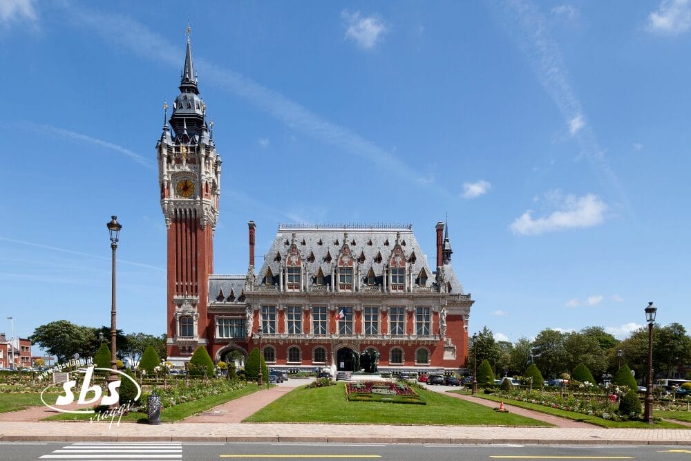 Un grande edificio storico in mattoni rossi con un'alta torre dell'orologio, come quelli che si trovano nelle capitali d'Europa come Londra o Parigi, si erge tra giardini curati sotto un cielo azzurro. Un sentiero conduce al grande ingresso.