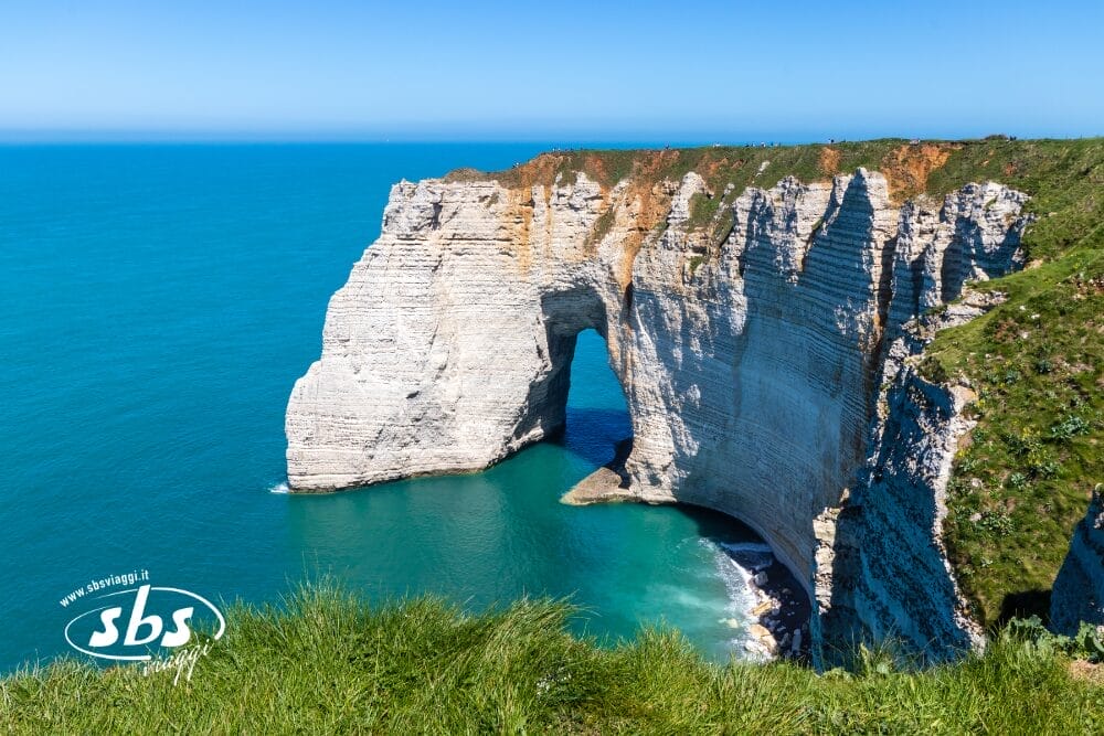 Scogliere di gesso bianco con un arco naturale si estendono in un mare turchese sotto un cielo azzurro e limpido, viste da un punto di vista erboso di bozza automatica. La scena è luminosa e costiera, con drammatiche formazioni rocciose.