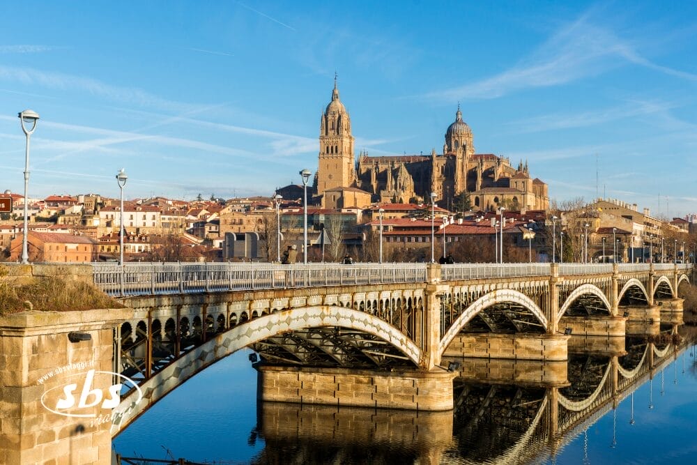Un ponte di pietra attraversa un fiume calmo con una cattedrale storica ornata e gli edifici della città sullo sfondo, sotto un cielo azzurro e limpido a Salamanca, in Spagna - una tappa iconica di qualsiasi tour Spagna Medievale de Le Meraviglie.