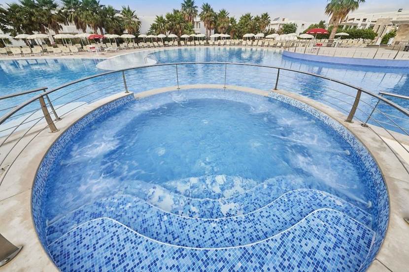 Una vasca idromassaggio rotonda e piastrellata con acqua gorgogliante è in primo piano e offre una vista automatica su una grande piscina all'aperto circondata da ombrelloni, sedie a sdraio e palme.