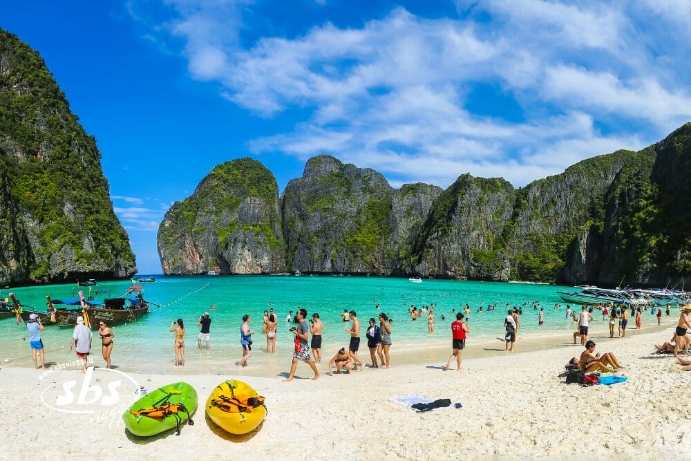 I turisti si godono una giornata di sole su una spiaggia tropicale in Thailandia con acqua turchese e scogliere verdi. La gente nuota, gioca e si rilassa sulla sabbia: l'ideale per un viaggio Thailandia o un Tour Thailandia da Nord a Phuket. Kayak e barche punteggiano la scena.