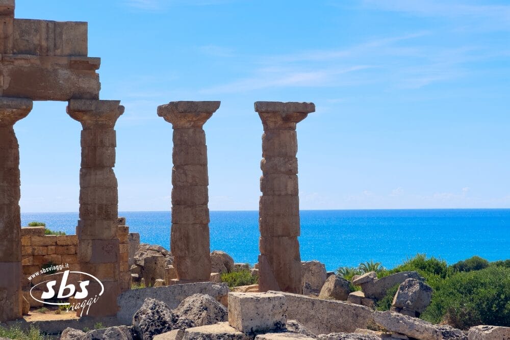 Antiche colonne di pietra e rovine si affacciano su un mare blu brillante sotto un cielo limpido. Il sito è circondato da una vegetazione verde, con la scritta "sbs viaggi" e la nota Bozza automatica nell'angolo in basso a sinistra.