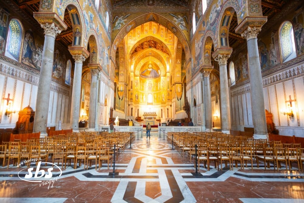 Interno di una grande cattedrale con ornati mosaici dorati, alti soffitti a volta, colonne di marmo e file di sedie di legno rivolte verso l'altare. Una persona si trova vicino alla navata centrale sotto le luci scintillanti, abbozzando i dettagli di una bozza automatica.