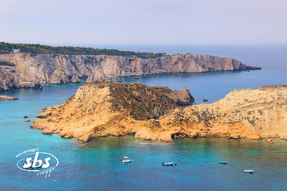 Un paesaggio costiero con scogliere rocciose e un'isola frastagliata di colore marrone chiaro circondata da acque limpide e blu. Alcune piccole imbarcazioni galleggiano nel mare e i pini coprono le scogliere lontane sotto un cielo nebuloso, come una Bozza automatica della natura.