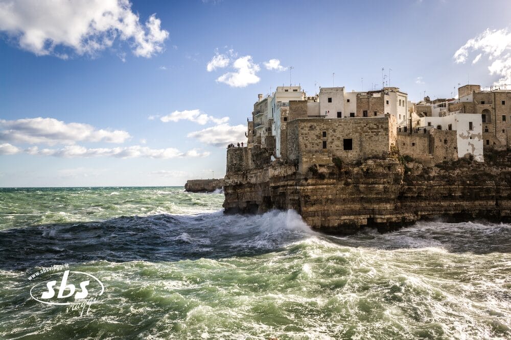Edifici bianchi si trovano in cima a una scogliera frastagliata, mentre le onde si infrangono contro le rocce sottostanti sotto un cielo parzialmente nuvoloso. Il mare turchese scintilla alla luce del sole, creando una bozza automatica mozzafiato di bellezza costiera.
