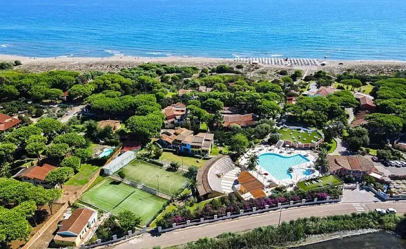 Vista aerea di un resort costiero con una grande piscina, campi da tennis, alberi verdi ed edifici vicino a una spiaggia sabbiosa e a un mare azzurro sotto un cielo limpido, perfetto per creare la vostra Bozza automatica.