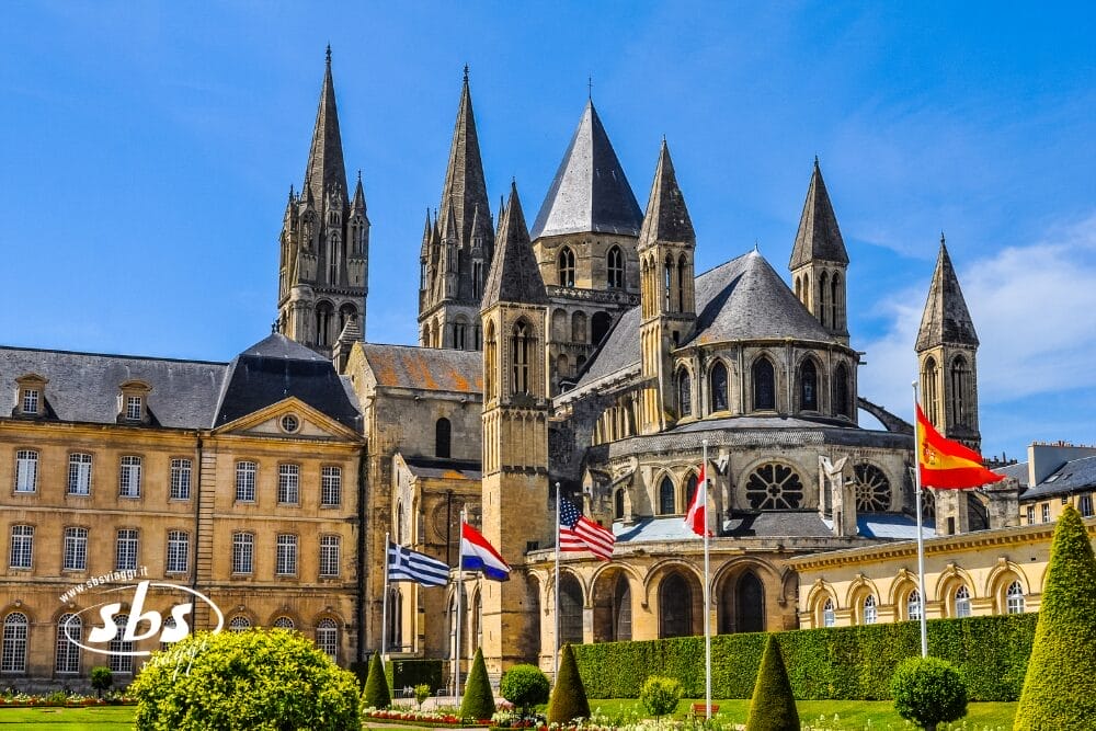 Storica abbazia in pietra con più guglie e grandi finestre ad arco, sullo sfondo di un cielo azzurro. In primo piano sono visibili diverse bandiere internazionali, siepi ben curate e un'insegna di Bozza automatica.