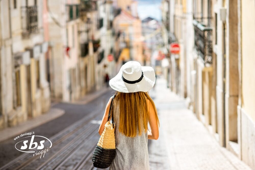 Una donna con i capelli lunghi, con un cappello da sole bianco e un vestito grigio, passeggia in una strada stretta e soleggiata, fiancheggiata da edifici. Porta una borsa di paglia lungo i binari del tram, mentre il logo "sbs viaggi" e Bozza automatica appaiono in un angolo.