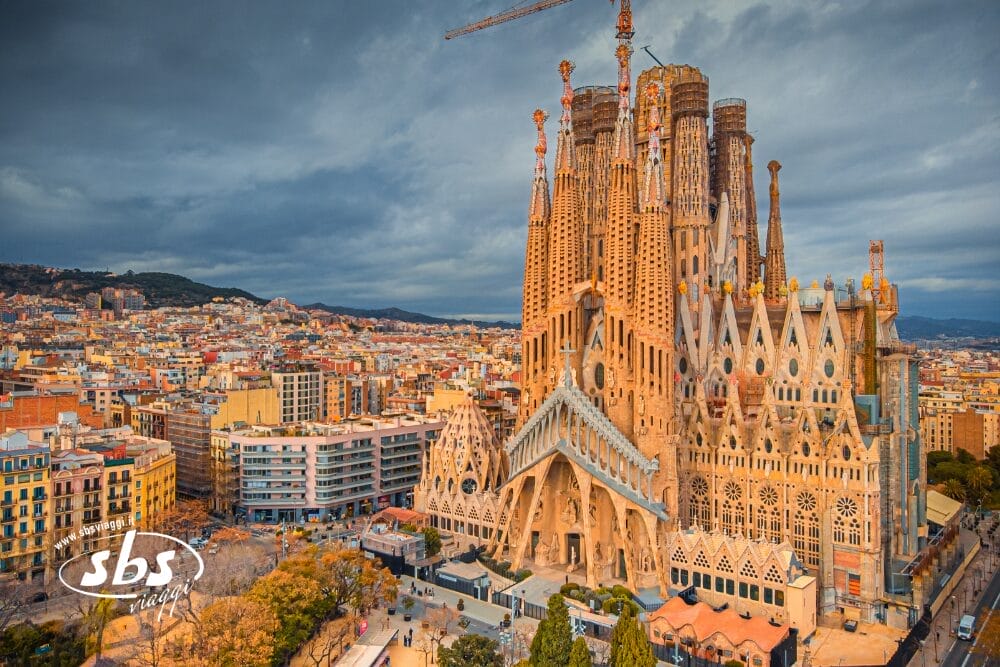 Veduta aerea della basilica della Sagrada Familia a Barcellona, in Spagna, con le sue alte torri e le gru che la sovrastano, come una Bozza automatica, circondata da edifici cittadini sotto un cielo nuvoloso.