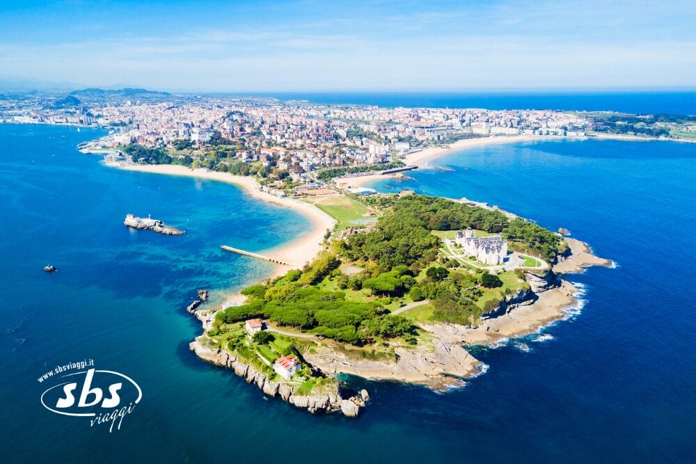 Veduta aerea di una penisola verde con un grande edificio circondato da alberi, che si estende nel mare blu, con una città e delle spiagge sullo sfondo sotto un cielo limpido. I testi "sbs viaggi" e "Bozza automatica" compaiono nell'angolo in basso a sinistra.