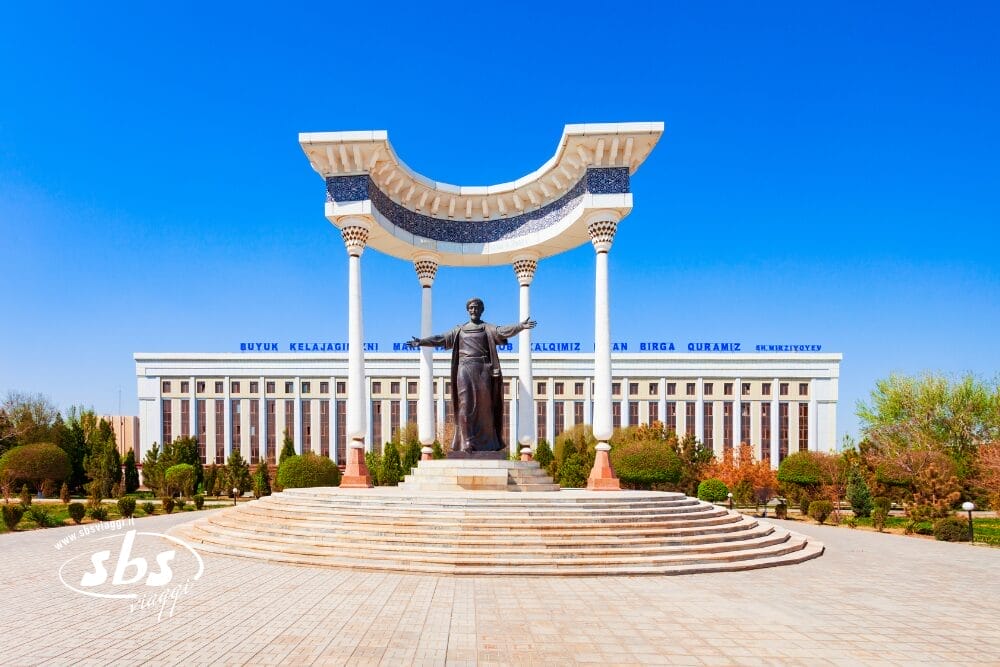 Una grande statua si erge sotto un arco decorativo bianco e blu in una piazza, con un edificio governativo sullo sfondo e alberi su entrambi i lati, il tutto sotto un cielo azzurro e limpido: una suggestiva Bozza automatica di eleganza urbana.