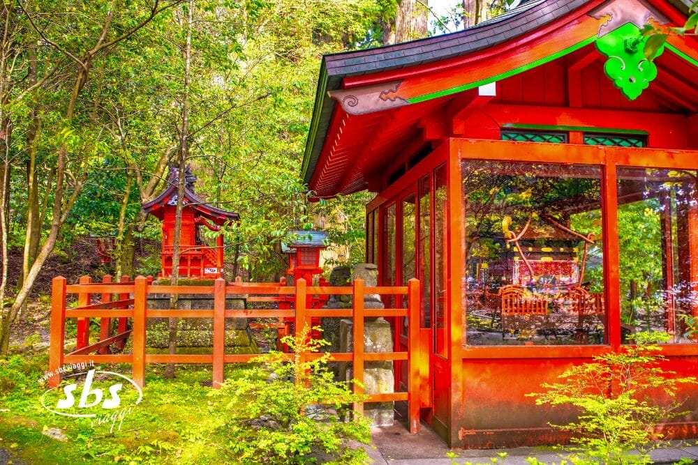 Un santuario giapponese di colore rosso vivo è circondato da alberi e piante verdi in Giappone, con la luce del sole che filtra attraverso il fogliame. Le superfici lucide del santuario riflettono il sereno ambiente naturale, perfetto per un tour in libertà.