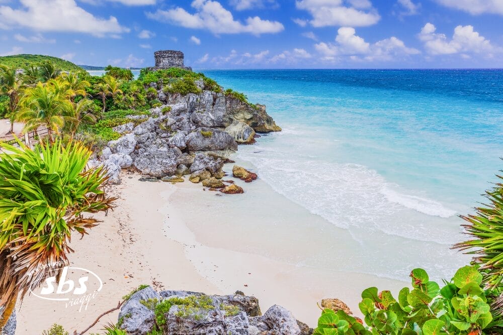 Spiaggia tropicale con acqua turchese, sabbia bianca e scogliere rocciose sormontate da una rovina Maya, circondata da palme e vegetazione lussureggiante sotto un cielo azzurro parzialmente nuvoloso a Tulum, Yucatán, Messico.
