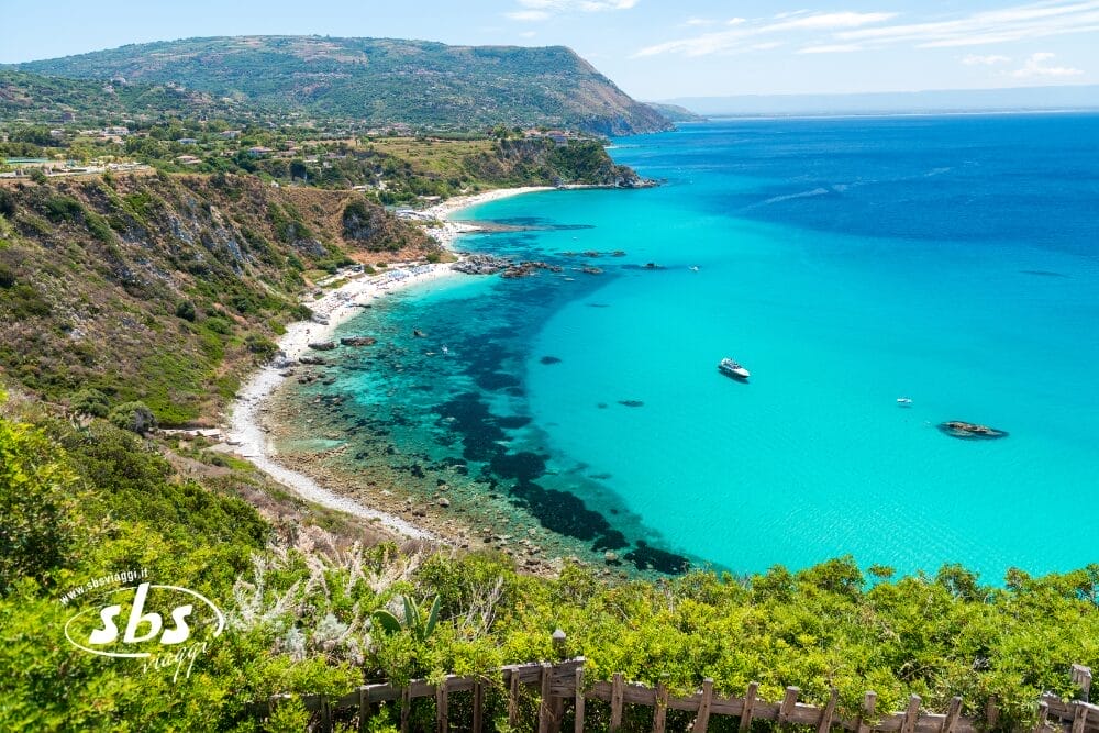 Una splendida vista costiera con acqua blu turchese, spiaggia sabbiosa, scogliere rocciose, vegetazione verde e barche ancorate vicino alla riva sotto un cielo limpido. Il logo Bozza automatica SBS Travel appare nell'angolo in basso a sinistra.